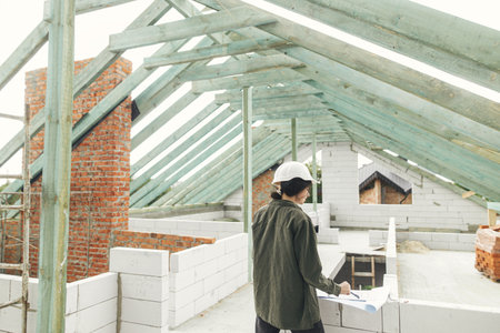 Young female architect with tablet checking blueprints against wooden roof framing of modern farmhouse. Stylish woman engineer in hard hat looking at digital plans at construction siteの写真素材