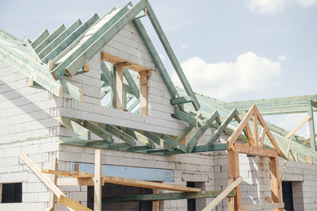 Unfinished modern farmhouse building. Wooden roof framing of mansard with dormer and aerated concrete block walls. Timber trusses, rafters and beams on blocks. New house constructionの写真素材