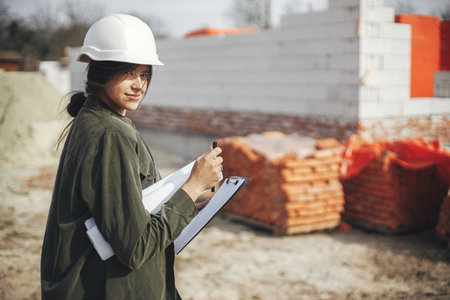 Young female engineer or construction worker in hardhat smiling against of building new modern house. Stylish happy woman architect with blueprints and plans at construction siteの写真素材