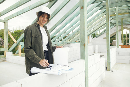 Young female engineer or construction worker in hardhat with plans at construction site. Stylish happy woman architect checking blueprints against wooden roof framing of modern farmhouseの写真素材