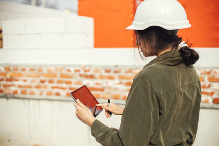 Stylish woman architect with tablet checking blueprints at construction site. Young female engineer or construction worker in hardhat looking at plans of new modern houseの写真素材