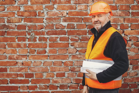 Senior male foreman in hardhat standing with blueprints at masonry brick wall at building new house. Portrait of happy man engineer or construction worker with plans at construction siteの写真素材