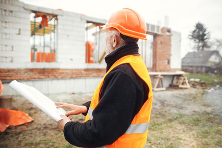 Senior male foreman in hardhat checking blueprints of building new modern house. Man engineer or construction worker looking at plans at construction site. Copy spaceの写真素材