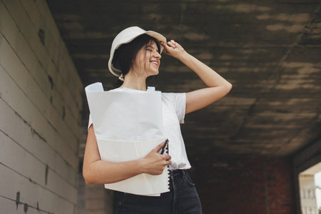 Young female engineer or construction worker in hardhat smiling against of building new modern house. Stylish happy woman architect with blueprints and plans at construction siteの写真素材
