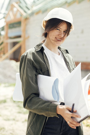 Stylish happy woman architect checking blueprints against modern farmhouse construction site. Young female engineer or construction worker in hardhat with plans of new modern houseの写真素材