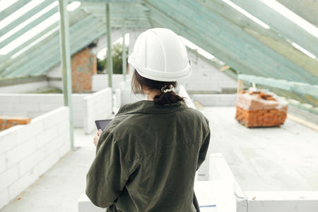 Stylish woman architect or engineer with tablet checking blueprints against wooden roof framing of modern farmhouse. Young female in hard hat looking at digital plans at construction siteの写真素材
