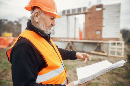 Senior man engineer or construction worker in hardhat looking at blueprints at building new modern house. Male architect or contractor checking plans at construction site. Copy spaceの写真素材