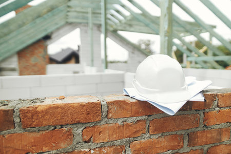 Architecture and House building concept. Hard hat with house blueprints on brick masonry on background of unfinished wooden roof framing. Safety helmet on construction site. Copy spaceの写真素材