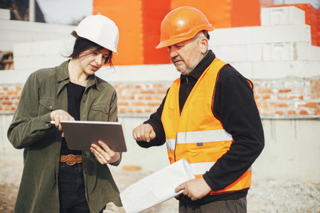 Stylish woman architect with tablet and senior foreman checking blueprints at construction site. Engineer and construction worker in hardhat looking at plans of new modern house. Teamworkの写真素材