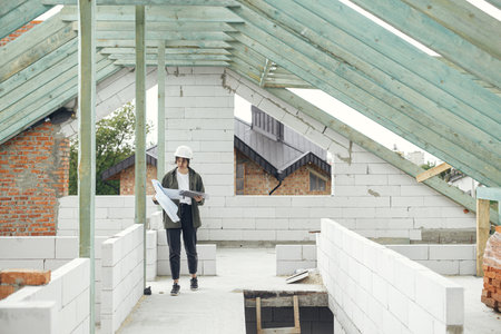 Stylish woman architect or engineer with tablet checking blueprints against wooden roof framing of modern farmhouse. Young female in hard hat looking at digital plans at construction siteの写真素材