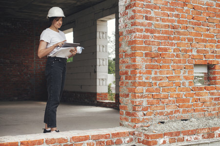 Stylish woman architect with tablet checking blueprints at construction site. Young female engineer or construction worker in hardhat looking at plans of new modern houseの写真素材