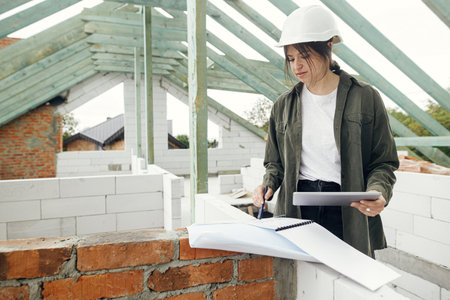 Young female architect with tablet checking blueprints against wooden roof framing of modern farmhouse. Stylish woman engineer in hard hat looking at digital plans at construction siteの写真素材