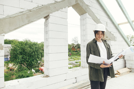 Young female engineer or construction worker in hardhat with plans at construction site. Stylish happy woman architect checking blueprints against wooden roof framing of modern farmhouseの写真素材