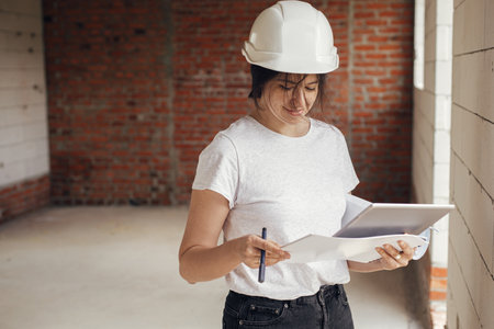 Young female architect with tablet checking blueprints against of building new modern house. Stylish happy woman engineer in hard hat looking at digital plans at construction siteの写真素材
