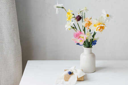 Stylish gift box with ribbon and spring flowers on rustic white table. Happy womens day and Mother's day. Beautiful daffodils and tulips gentle bouquet and present. Space for textの写真素材