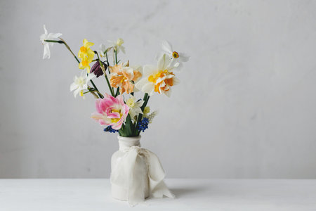 Stylish spring flowers in vase with ribbon on rustic white table. Happy womens day and Mother's day. Beautiful daffodils and tulips gentle bouquet still life. Space for textの写真素材