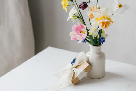 Happy womens day and Mother's day. Stylish gift box with ribbon and spring flowers on rustic white table. Beautiful daffodils and tulips gentle bouquet and present. Space for textの写真素材