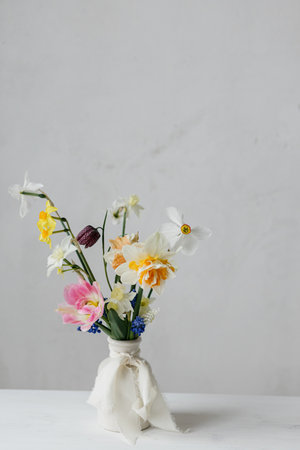 Stylish spring flowers in vase with ribbon on rustic white table. Happy womens day and Mother's day. Beautiful daffodils and tulips gentle bouquet still life. Space for textの写真素材