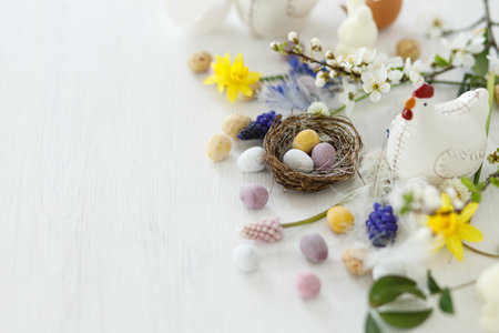 Happy Easter! Stylish easter chocolate eggs in nest, spring flowers, chicken figurine on white rustic wooden table. Easter modern simple banner, space for text. Seasons greetingsの写真素材
