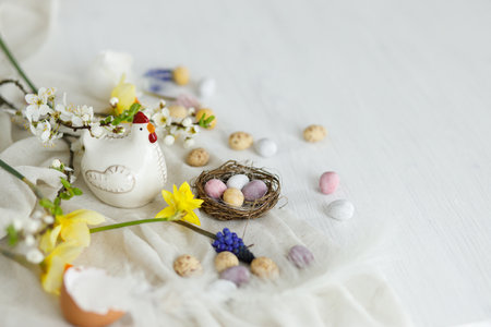 Happy Easter! Stylish easter chocolate eggs in nest, spring flowers, chicken figurine on white rustic wooden table. Easter modern simple banner, space for text. Seasons greetingsの写真素材