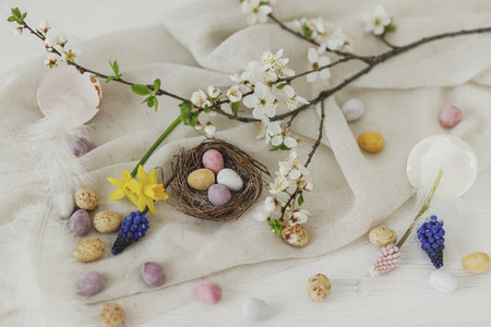 Happy Easter! Stylish easter chocolate eggs in nest, spring flowers, feathers and linen cloth on white rustic wooden table. Easter modern simple banner, space for text. Seasons greetingsの写真素材