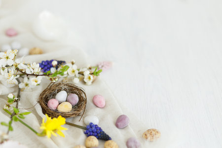 Colorful easter chocolate eggs in nest, spring flowers, feathers and linen border composition on white wooden table. Space for text. Happy Easter! Seasons greetingsの写真素材