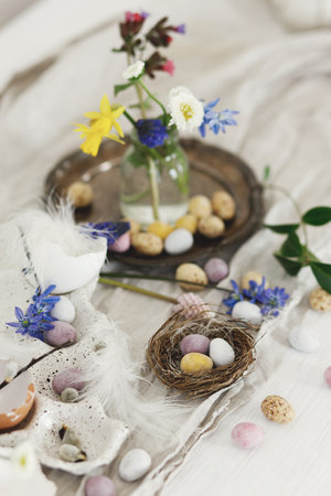 Stylish easter chocolate eggs in nest, spring flowers, feathers and linen cloth on rustic wooden table. Easter modern simple decoration still life. Happy Easter!の写真素材
