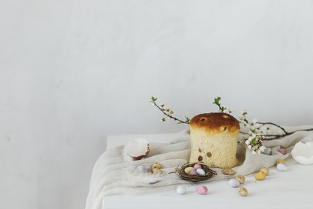 Colorful easter chocolate eggs in nest, spring flowers, traditional bread and linen cloth on rustic white wooden table. Space for text. Happy Easter! Seasons greetingsの写真素材