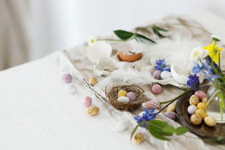 Happy Easter! Colorful easter chocolate eggs in nest, spring flowers, feathers and linen cloth on rustic wooden table. Space for text. Easter modern simple decorationの写真素材