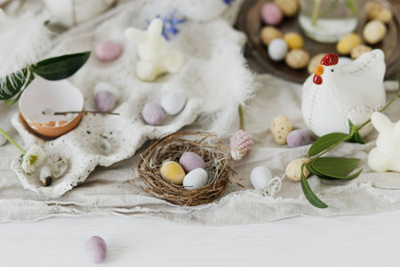 Colorful easter chocolate eggs in nest, spring flowers, chicken figurine and linen cloth on rustic wooden table. Space for text. Easter modern simple decoration. Happy Easter!の写真素材