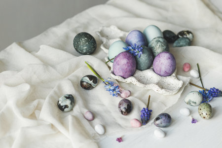 Happy Easter! Stylish easter eggs in modern tray and spring flowers on linen cloth on rustic white table. Easter simple banner, space for text. Natural painted marble and purple eggs still lifeの写真素材