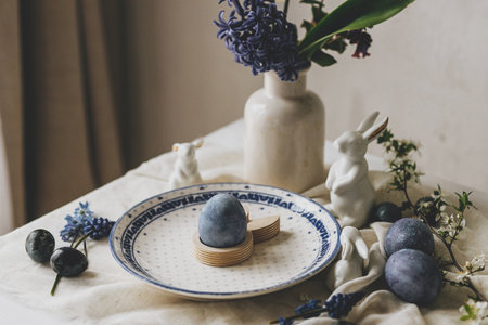 Happy Easter! Stylish easter egg in bunny figurine on vintage plate and spring flowers on linen, rustic table setting. Natural painted blue eggs and hyacinth blooms. Modern minimal still lifeの写真素材