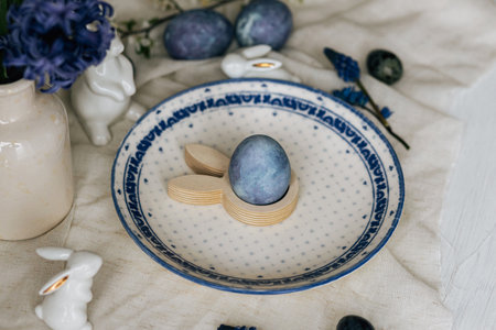 Happy Easter! Stylish easter egg in bunny figurine on vintage plate and spring flowers on linen, rustic table setting. Natural painted blue eggs and hyacinth blooms. Modern minimal still lifeの写真素材