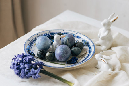 Stylish easter eggs on vintage plate, bunny and spring flowers on rustic table. Happy Easter! Natural dye blue eggs, purple hiacynt blossoms on linen napkin. Holiday setting, minimal still lifeの写真素材