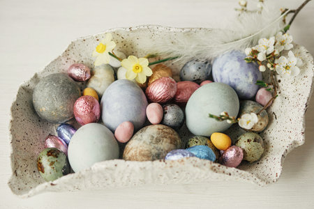 Stylish easter eggs in modern bowl with cherry blossom and daffodils composition on rustic table....
