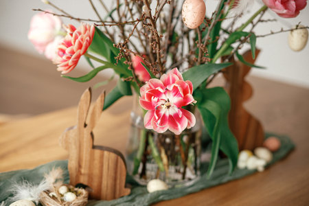 Stylish easter decoration on wooden table close up in room. Beautiful tulips bouquet with willow branches, easter eggs and wooden bunny figurines decor. Happy Easter!の写真素材
