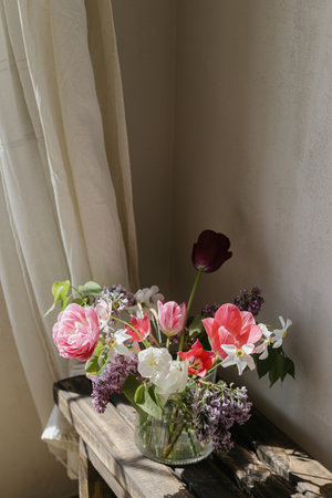Stylish spring bouquet on rustic wooden bench. Beautiful lilacs, tulips and daffodils in glass vase on background of grey wall and linen curtains. Floral still life. Happy Mothers dayの写真素材