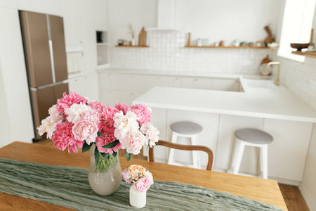 Modern kitchen interior. Beautiful peonies in vase on wooden table on background of stylish white kitchen with appliances in new scandinavian house.Summer floral arrangementの写真素材