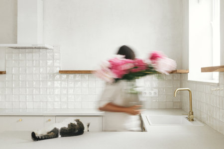 Blurred woman with her cat arranging peonies in vase on background of minimal white kitchen in new modern home. Motion image of young female decorating house after renovation, housekeepingの写真素材