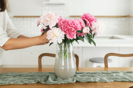 Woman arranging beautiful peonies in vase in new modern home. Young female decorating house with flowers on background of minimal white kitchen, hands with flowers close upの写真素材