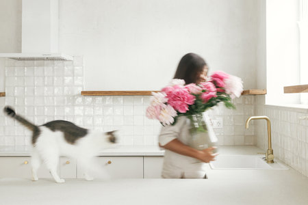 Blurred woman with her cat arranging peonies in vase on background of minimal white kitchen in new modern home. Motion image of young female decorating house after renovation, housekeepingの写真素材