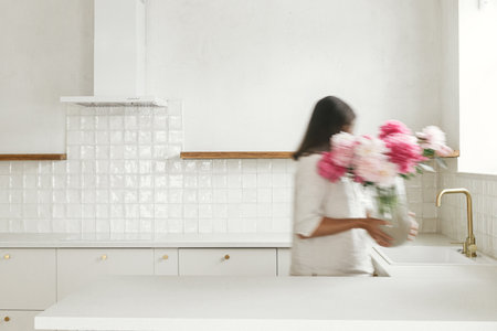 Blurred woman arranging peonies in vase on background of minimal white kitchen in new modern home. Motion image of young female decorating house after renovation, housekeepingの写真素材