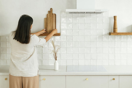 Stylish woman unpacking and decorating shelves with utensils and cutting boards in new minimal white kitchen. Housewife cleaning up kitchen after moving  in new modern scandinavian homeの写真素材