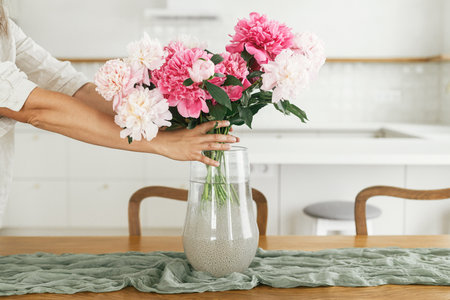 Woman arranging beautiful peonies in vase on wooden table in new modern home. Young female decorating dining room with flowers on background of minimal white kitchen, moving inの写真素材