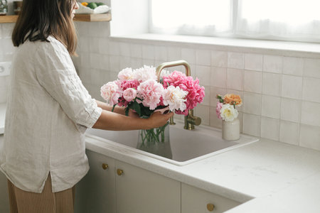Woman arranging beautiful peonies in vase at sink with brass faucet and granite countertop. Florist holding pink peony and scissors in new modern kitchen, hands with flowers close upの写真素材