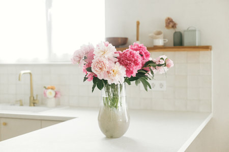Beautiful peonies in vase on granite countertop on background of stylish white kitchen with wooden shelves and appliances in new house. Modern minimal kitchen interiorの写真素材