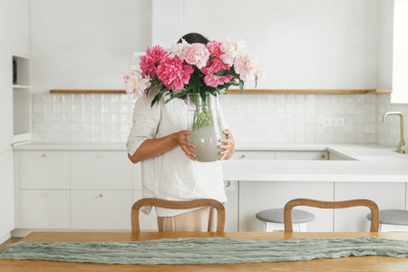 Woman holding beautiful peonies in vase at wooden table in new modern home. Young female decorating dining room with flowers on background of minimal white kitchen, moving in and housekeepingの写真素材