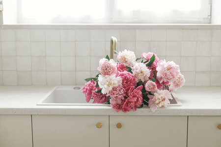 Beautiful peonies in sink on background of brass faucet and white counter in new scandinavian house. Pink peony flowers in modern kitchen interior, summer floral arrangementの写真素材