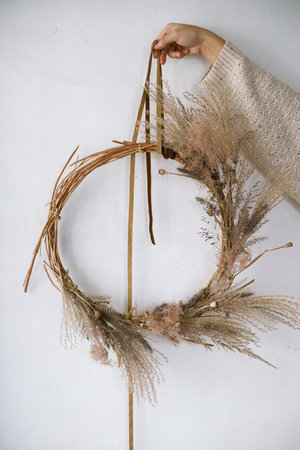 Hands holding wreath with dried grass, herbs, leaves, ribbon at white wall. Woman making stylish autumn wreath. Fall decor and arrangement in home.の写真素材