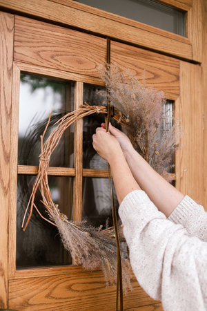 Woman decorating front door with stylish autumn wreath. Rustic wreath with dried grass, herbs and ribbon in hands. Fall decor of house entrance. Thanksgiving and Halloweenの写真素材